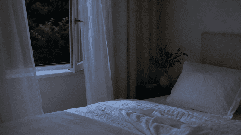 A cool, dark, minimalist bedroom with a slightly open window, light curtains, and white linen bedding suggesting an ideal sleep temperature environment