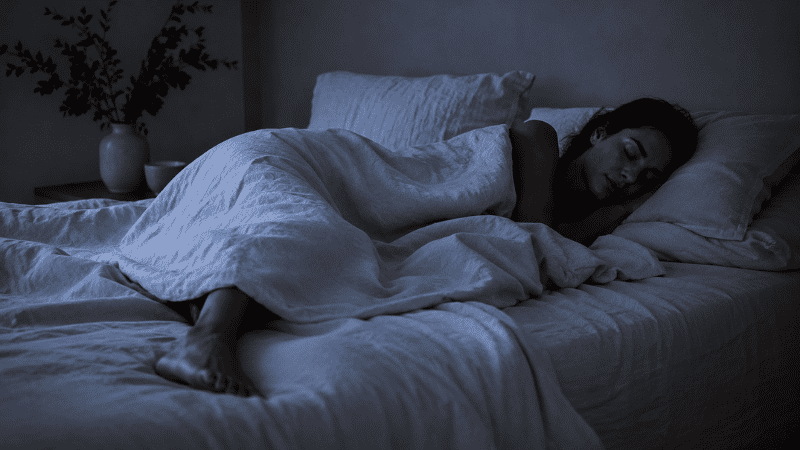 A person sleeping peacefully in a cool dark bedroom with light linen bedding, one foot slightly outside the covers, representing deep restful sleep in an optimized temperature environment