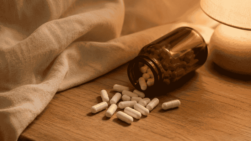 Magnesium glycinate capsules in an amber glass bottle on a wooden nightstand beside a warm lamp, representing a natural sleep supplement