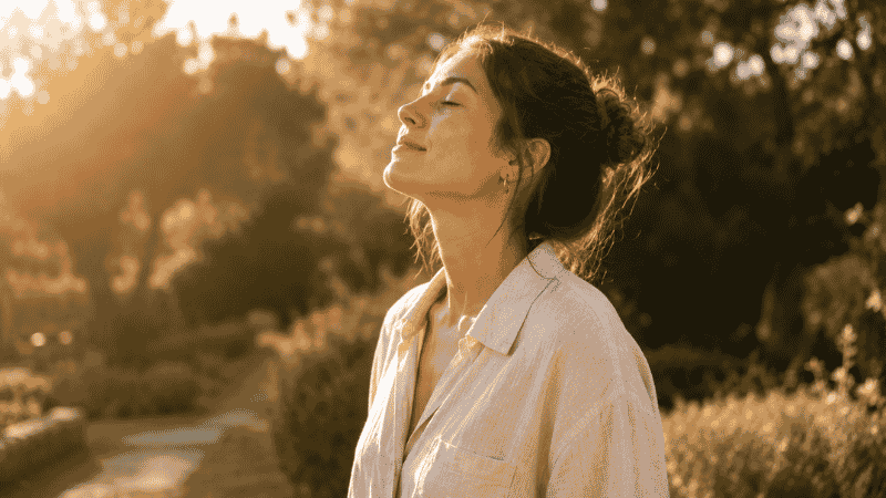 A person standing outdoors in soft golden morning sunlight with eyes closed and face lifted toward the light, representing morning light exposure for circadian rhythm reset