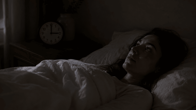 A person lying awake in a dark bedroom at 3am, staring at the ceiling with a dim clock visible on the nightstand, representing middle-of-the-night insomnia