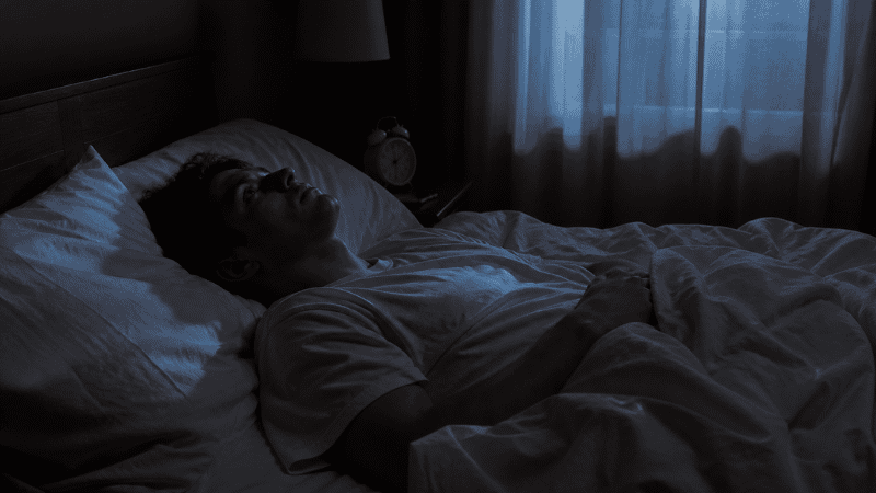 Person lying awake in bed at night staring at the ceiling, unable to fall asleep