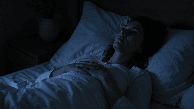 A person lying awake in a dark bedroom at night with their eyes open and hands resting on their chest, looking restless, representing anxiety-driven insomnia and difficulty sleeping with anxiety