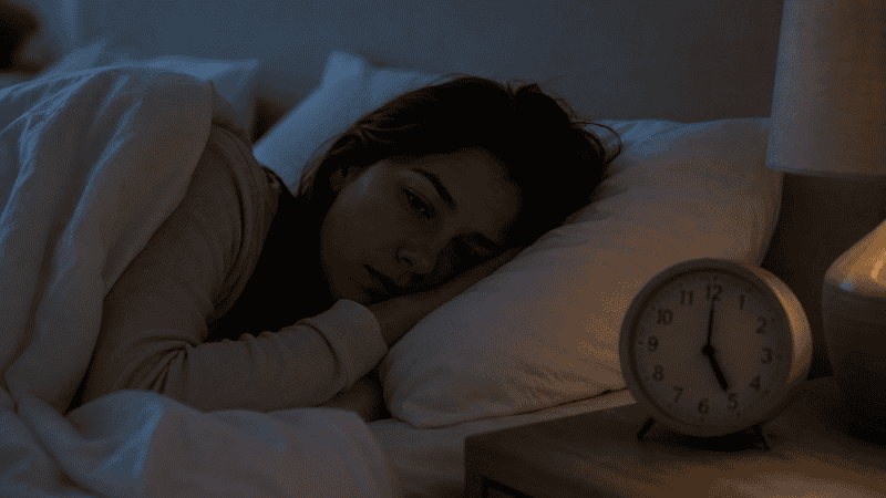 A person lying in bed in the early morning with eyes half open and an alarm clock showing 5am, looking tired and unrested, representing the effects of only 5 hours of sleep