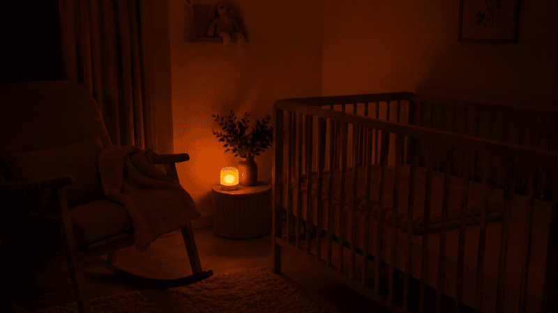 A dimly lit nursery at night with only a soft red night light glowing, representing low-stimulation lighting during nighttime feeds to preserve melatonin and support postpartum sleep