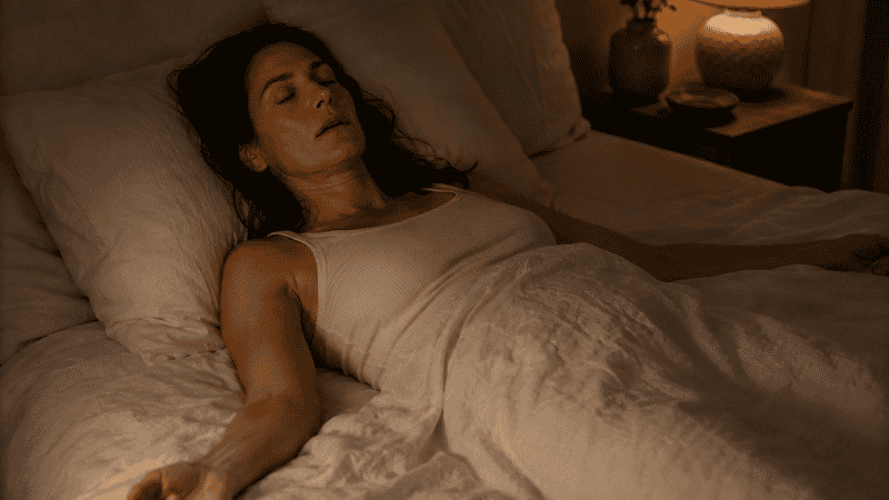 A person lying in bed with shoulders visibly relaxed and arms loose at their sides, representing the muscle release phase of progressive muscle relaxation for sleep