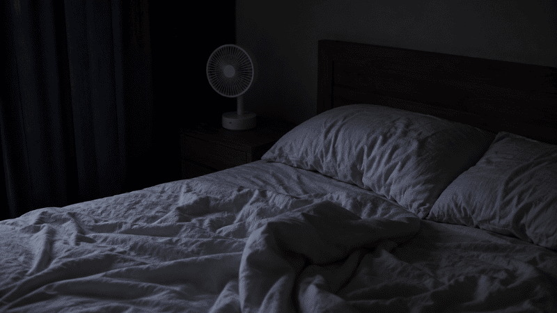 A bedroom at night with blackout curtains, a small fan, and dim warm lamp, representing the key environmental factors — temperature, light, and sound — that affect waking in the middle of the night