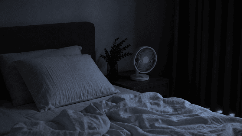 A cool dark minimal bedroom at night with blackout curtains, a small bedside fan, and light breathable linen bedding, representing an optimized sleep environment for sleeping through the night