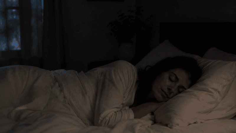 A person sleeping peacefully and undisturbed through the night in a dark calm bedroom with white linen bedding, representing the goal of sleeping through the night without waking