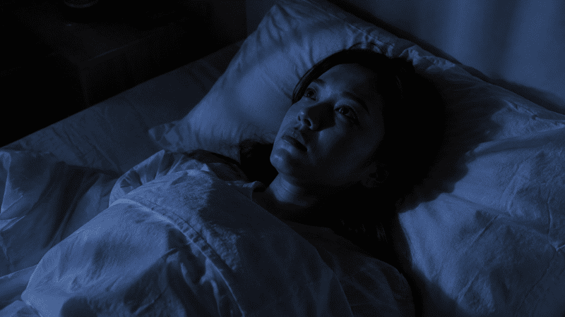 A person lying awake in bed at night with eyes open and a tense expression, representing the neurological connection between lack of sleep and anxiety