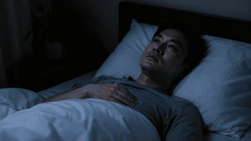 A person lying awake in bed at night looking tense with one hand on their chest, representing elevated cortisol and sympathetic nervous system activation that disrupts the nocturnal blood pressure dip