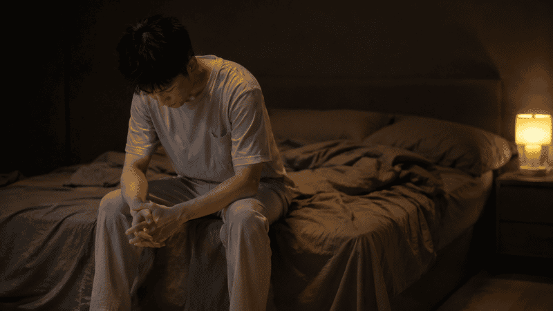 A person sitting on the edge of a bed in a dim bedroom at night with their head slightly bowed, representing the experience of nighttime anxiety and searching for calm