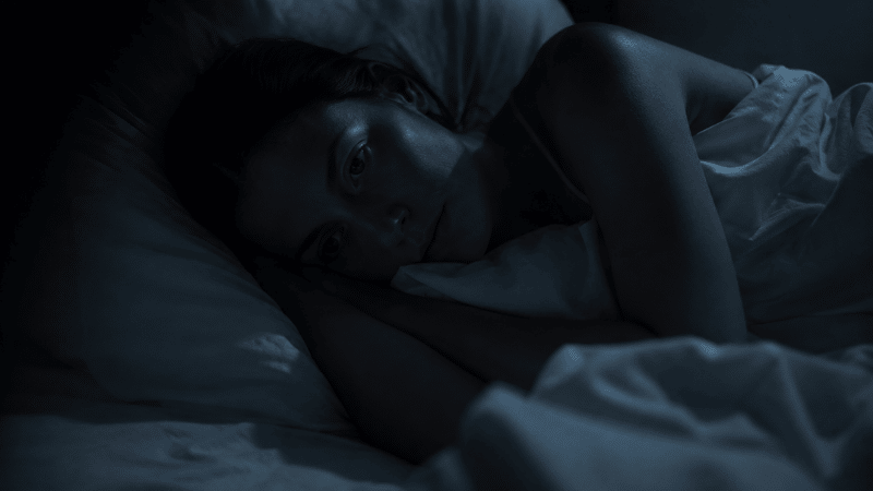 A person lying awake in a dark bedroom at night with their eyes open, looking quietly restless, representing the experience of nighttime anxiety when the house goes quiet